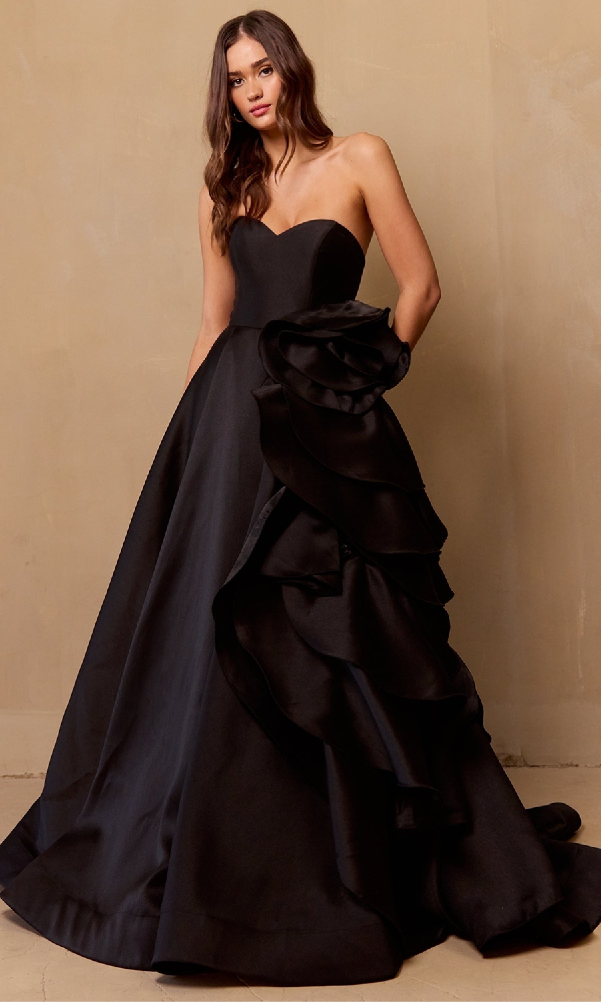 Woman wearing the Amelia Couture SU085 Strapless Prom Ball Gown, a black dress with a fitted bodice and full skirt adorned with dramatic ruffled detailing, standing against a beige wall.